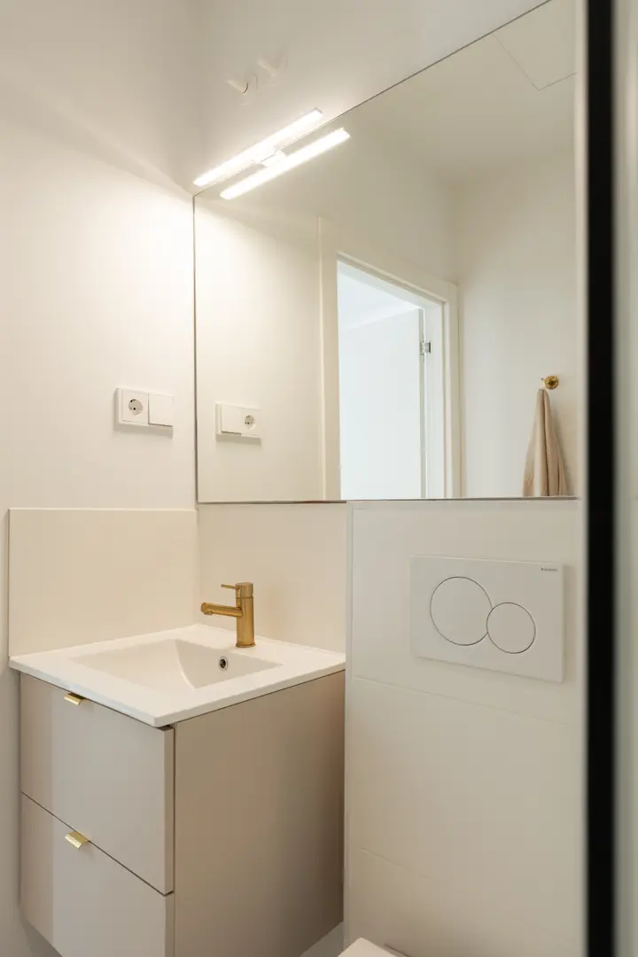 Compact bathroom in the Toro residence featuring a beige vanity unit with a gold-coloured tap and a large, illuminated mirror