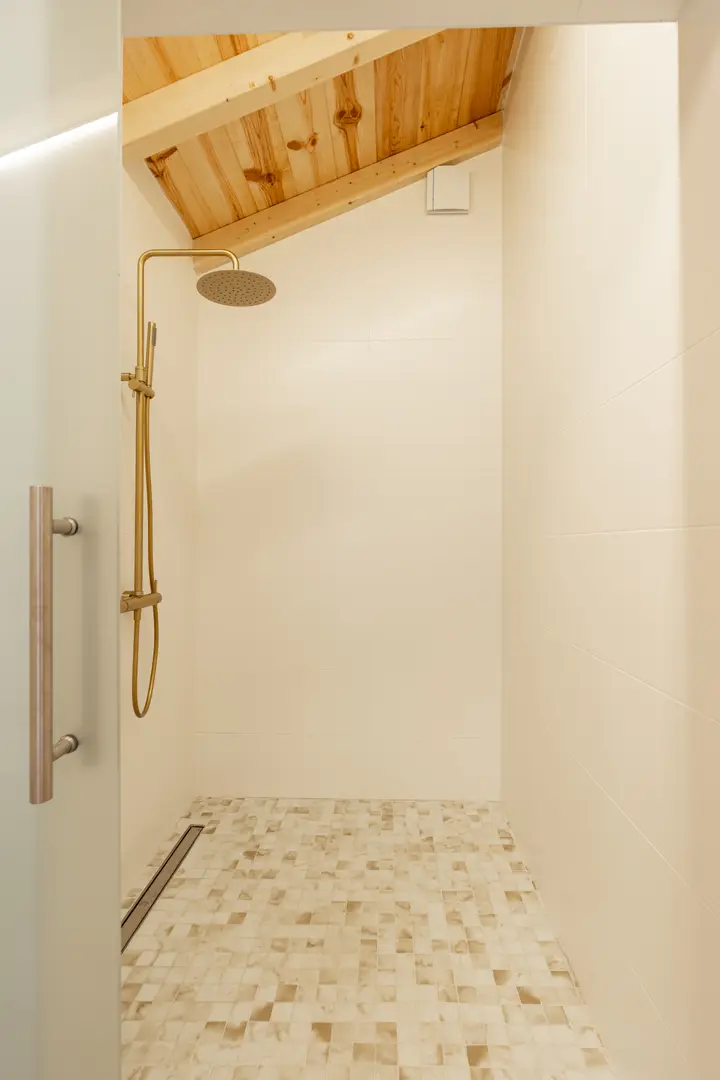 A walk-in shower in the Toro residence with gold-coloured fittings, mottled floor tiles, and an exposed pine beam ceiling