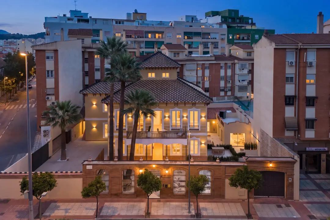 Aerial view of the illuminated Palacete 10 villa and its grounds at dusk, surrounded by palm trees and city buildings in Málaga