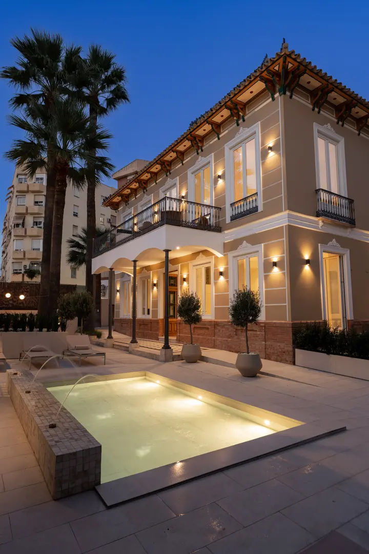 The illuminated facade of Palacete 10 at dusk, seen from the courtyard with its pond and palm trees