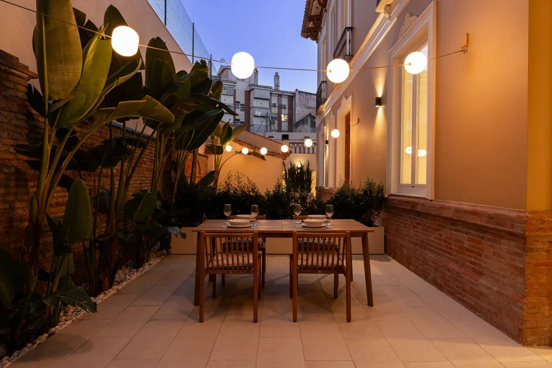 A wooden dining table set for four on a paved courtyard at dusk, lit by strings of globe lights