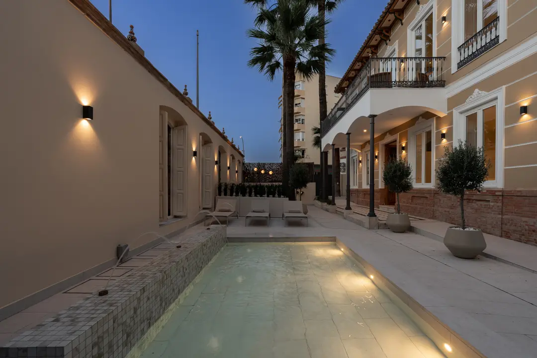 The illuminated courtyard of Palacete 10 at dusk, with its long pond and fountains, sun loungers, and the historic villa