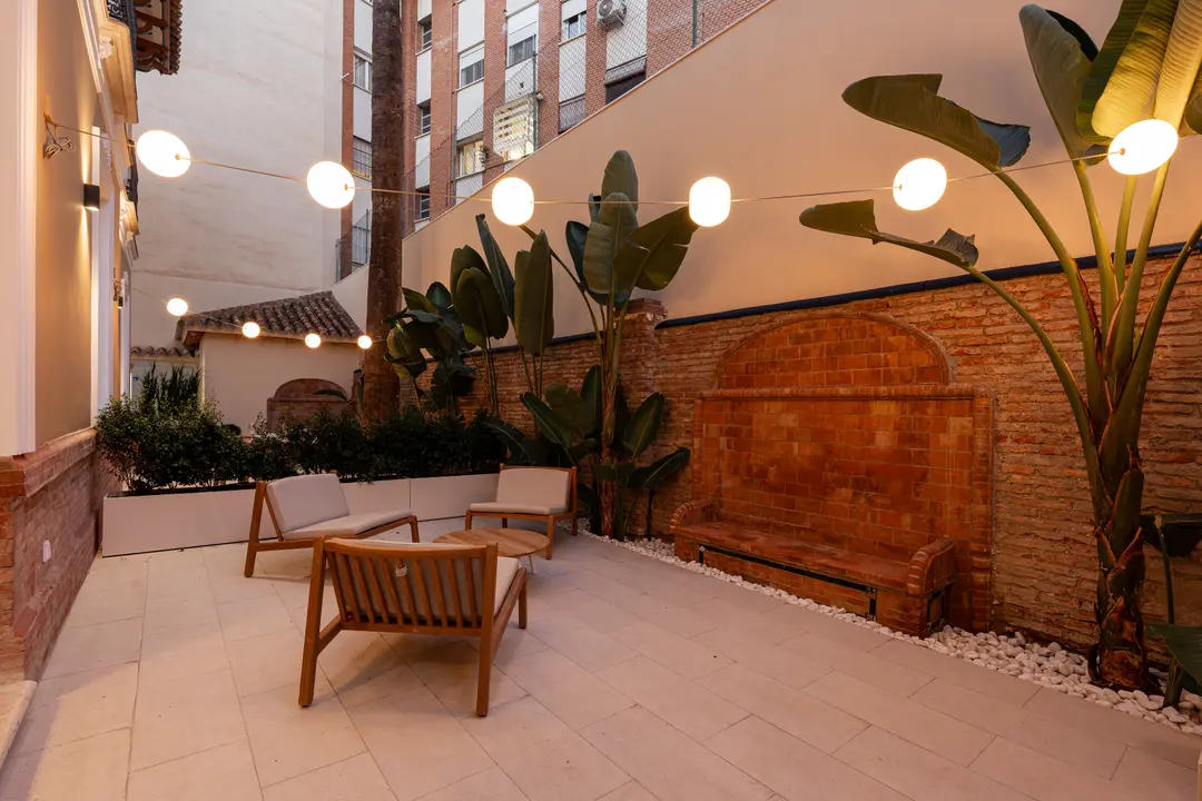 The courtyard at Palacete 10 at dusk, with wooden outdoor furniture, a tiled bench, tropical plants and warm globe lights