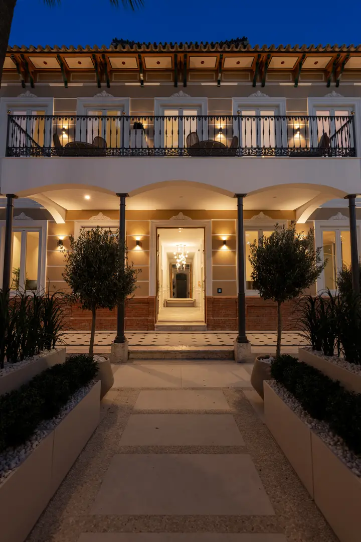 The illuminated facade of Palacete 10 at dusk, viewed from the garden path leading to the open entrance door