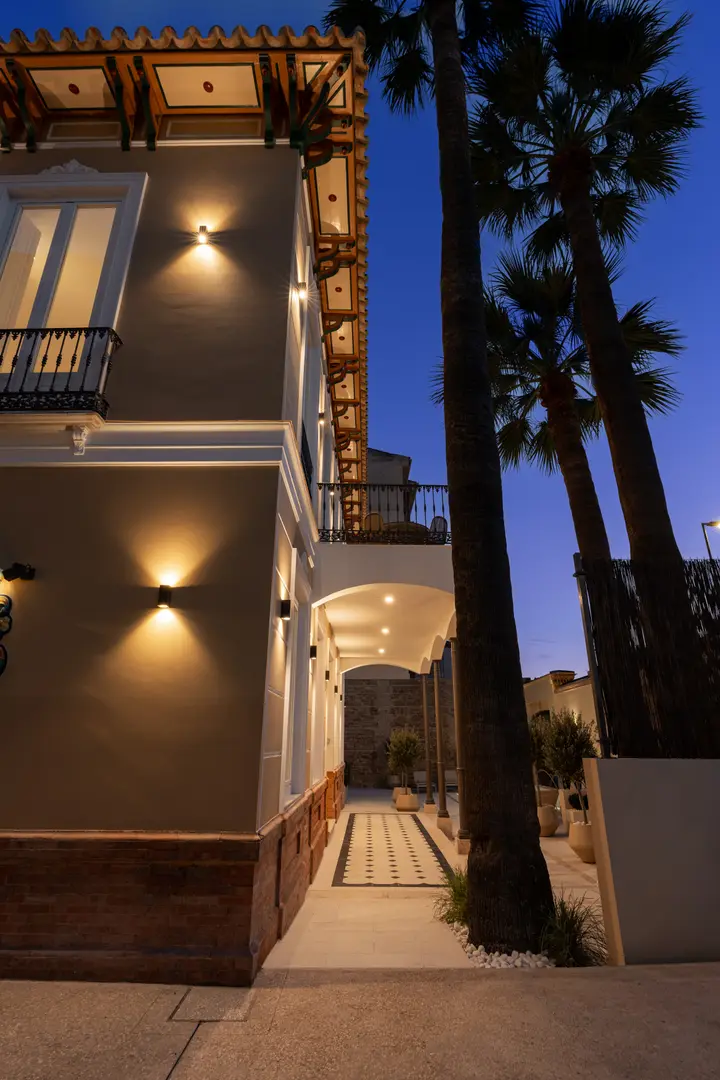 The restored facade of Palacete 10 at dusk, with warm lighting, a covered walkway, and tall palm trees against a deep blue sky