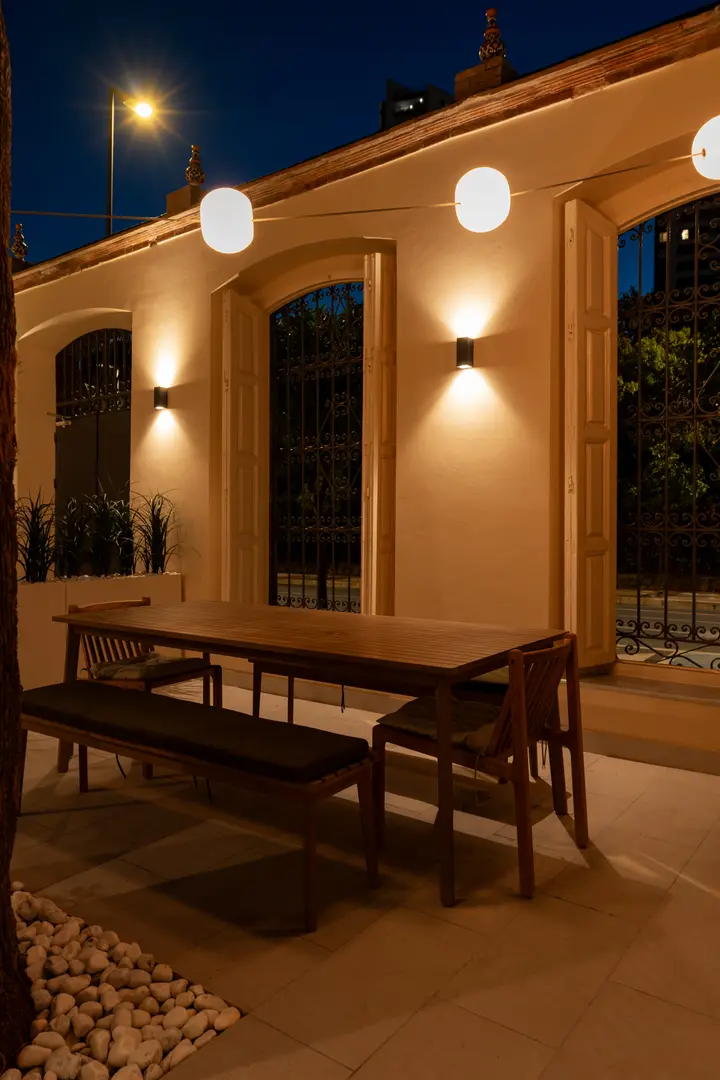 A large wooden dining table and bench on a stone patio at night, warmly lit by wall sconces and a string of globe lights