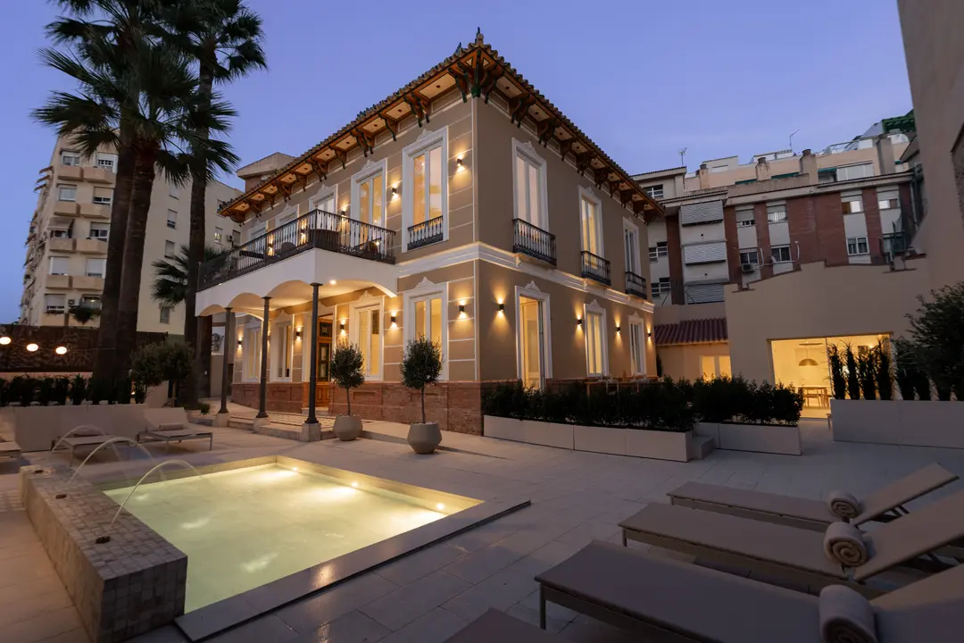 The restored Palacete 10 villa and its courtyard at dusk, with the illuminated pond, sun loungers, and tall palm trees