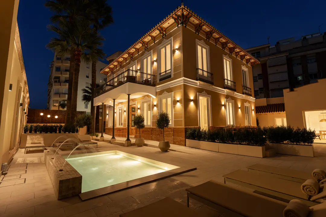 The ochre facade of Palacete 10 illuminated at night, seen from the courtyard with its pond, fountains, and sun loungers