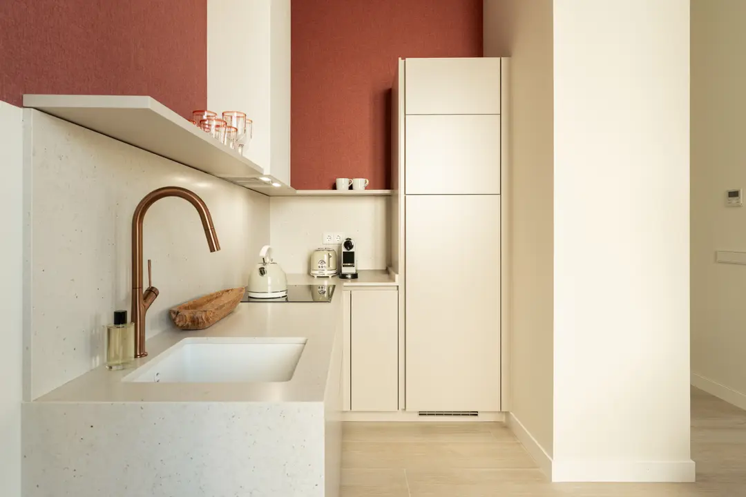The compact kitchen in the Vino residence, with cream units, a stone worktop, copper tap, and a dark red wall