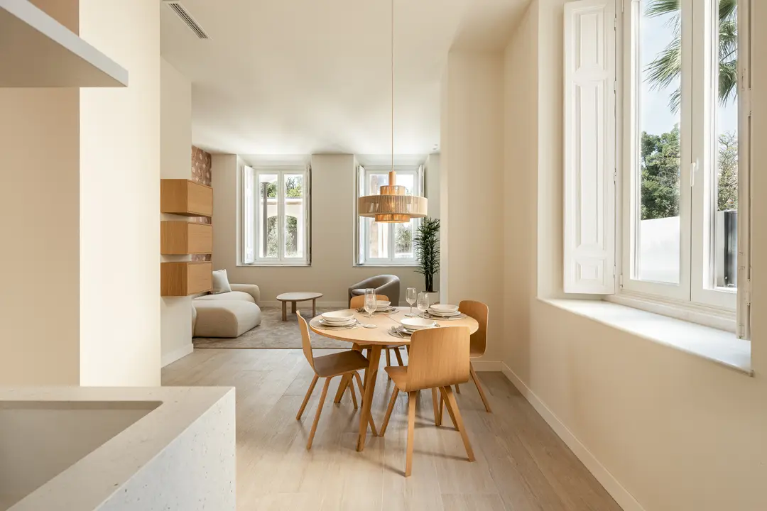 A round wooden dining table under a woven pendant lamp, with the bright, open-plan living room of the Vino residence behind