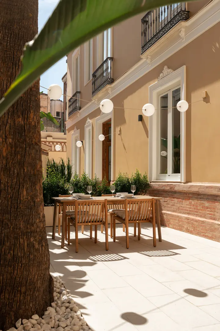 A wooden dining table set for six on the sunny private patio of the Vino residence, seen from behind a palm tree