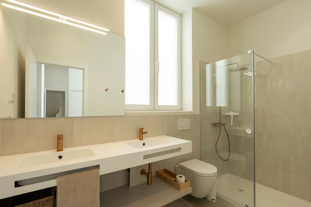 The bathroom in the Vino residence features a double vanity with brushed gold taps, a walk-in rain shower, and neutral-toned tiles