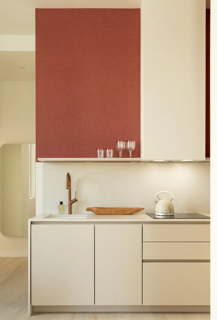 Detail of the kitchen in the Vino residence, showing off-white cabinets, a stone worktop, and a copper tap against a wine-red wall