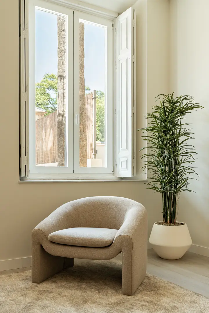 A sunlit corner in the Vino residence, with a modern beige armchair and a tall plant beside a window