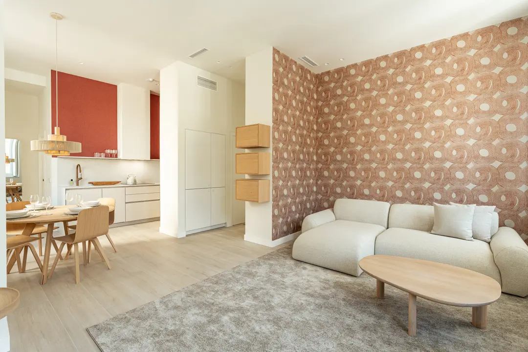 Bright open-plan living room in the Vino residence, with a cream sofa, red patterned wallpaper, and a dining area and kitchen