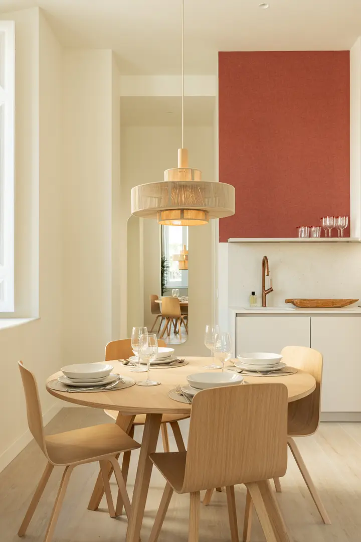 A round wooden dining table set for four under a woven pendant lamp in the Vino residence's bright, open-plan space