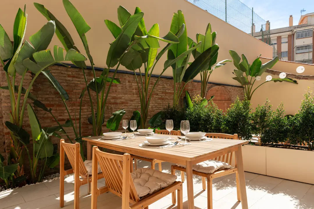 The sunny private terrace of the Vino residence, with a wooden dining table set for six against a brick wall with lush plants