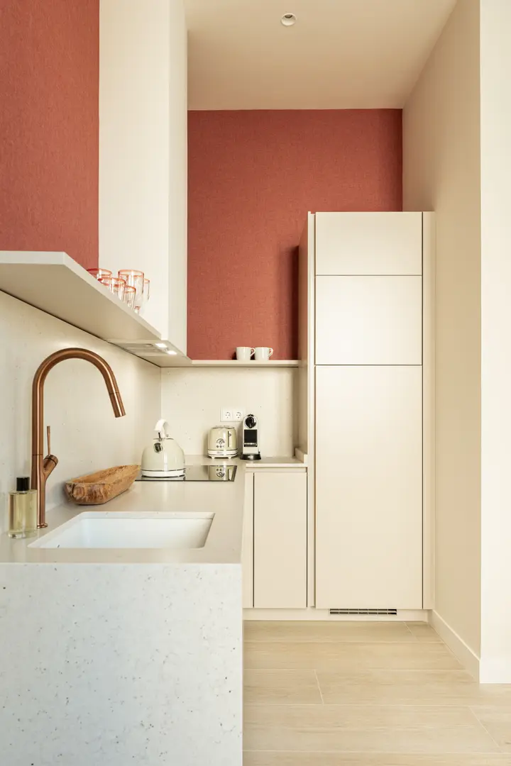 The compact kitchenette in the Vino residence, with cream cabinets, a speckled stone worktop, and a copper tap against terracotta-red walls