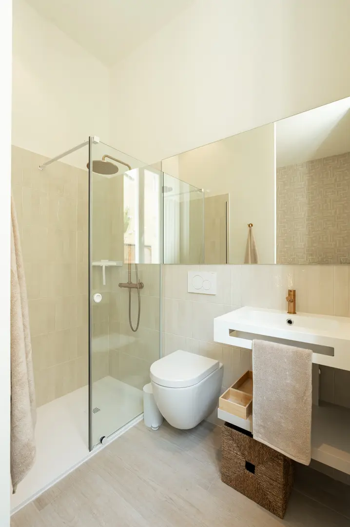 The bathroom in the Vino residence, with beige tiles, a glass walk-in shower, and a modern white console sink