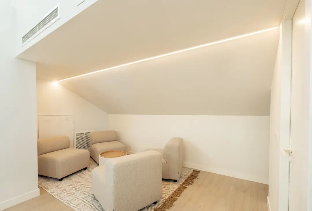 A lounge area in the attic work suite, with low beige armchairs arranged on a rug under a sloped ceiling