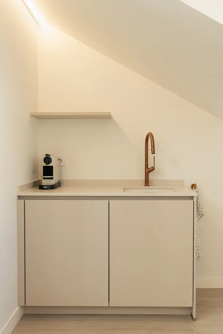 The kitchenette in the attic work suite, with beige cabinets, a stone worktop, and a modern copper tap