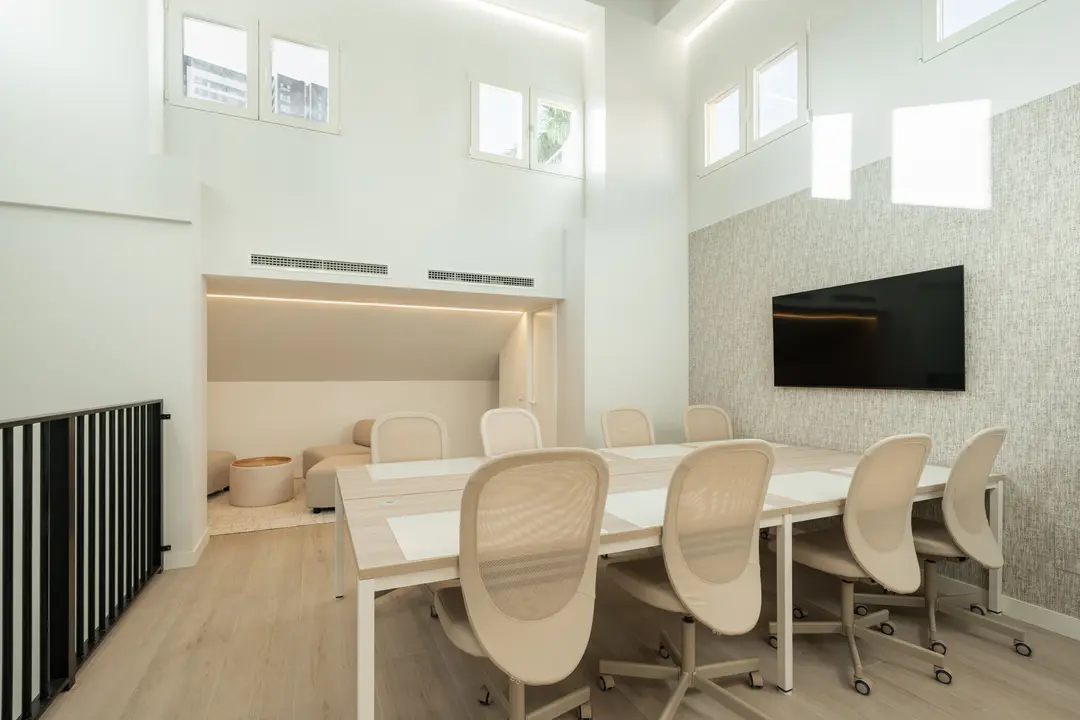 The bright attic work suite with a long meeting table, wheeled chairs, a wall-mounted screen, and a breakout nook